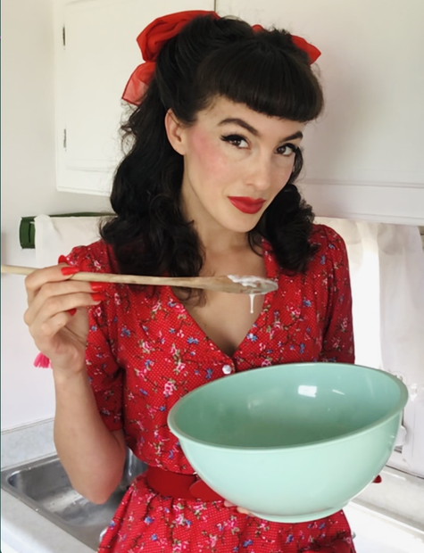 Sugar May holds a mixing bowl and wooden spoon. They are wearing a red dress and have black hair styled in 1940s victory rolls with a red bow