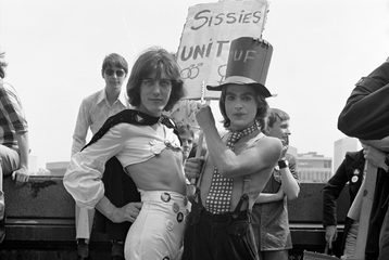 Two people in 1976, wearing non-binary clothes and carrying a sign that says 'Sissies Unite'
